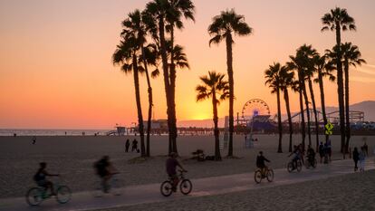 Gente andando en bicicleta por el sendero frente a la playa al atardecer en Santa Mónica, en California. Un bosque de palmeras se recorta contra la puesta de sol. El muelle de Santa Mónica y la noria se ven al fondo.