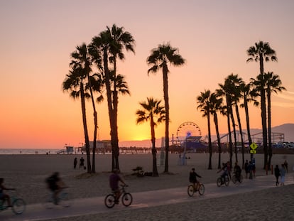 Gente andando en bicicleta por el sendero frente a la playa al atardecer en Santa Mónica, en California. Un bosque de palmeras se recorta contra la puesta de sol. El muelle de Santa Mónica y la noria se ven al fondo.