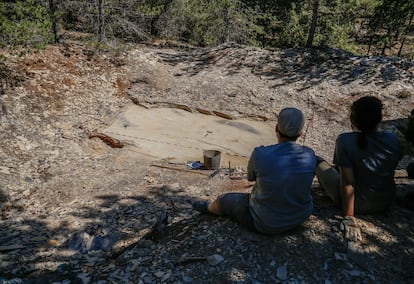 Dos estudiantes descansan frente a la Cata Botánicos mientras el dron sobrevuela la superficie descubierta para documentarla. La sombra del aparato se aprecia en la izquierda superior del perímetro de estudio. 