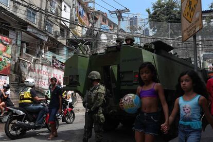 Duas meninas caminham ao lado de uma viatura militar.