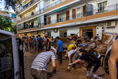 Un grupo numeroso de voluntarios trabajan en la limpieza de una calle en Alfafar el 2 de noviembre. El agua alcanzó una altura de hasta dos metros, inundando por completo las plantas bajas de los edificios. Una señora mayor observa la escena desde el balcón de su casa en la primera planta. La imagen es del 2 de noviembre, solo cuatro días después de las devastadoras inundaciones. El esfuerzo colectivo de los vecinos y voluntarios para limpiar las calles de barro y escombros destaca ante la ausencia de ayuda gubernamental, una situación que incrementa la sensación de abandono entre los afectados.