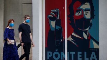 Dos personas pasean por delante de un graffiti con la imagen del director del centro de Coordinación de Alertas y Emergencias Sanitarias, Fernando Simón, en la puerta de un comercio de Barcelona