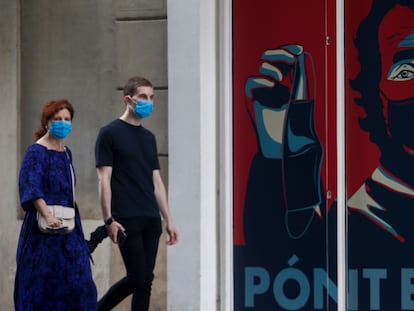 Dos personas pasean por delante de un graffiti con la imagen del director del centro de Coordinación de Alertas y Emergencias Sanitarias, Fernando Simón, en la puerta de un comercio de Barcelona