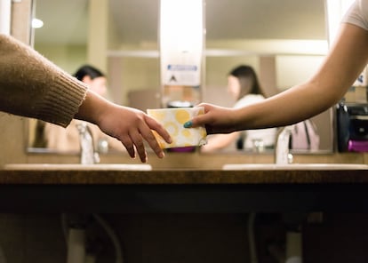 Uma pessoa entrega um absorvente a outra num ambiente escolar.