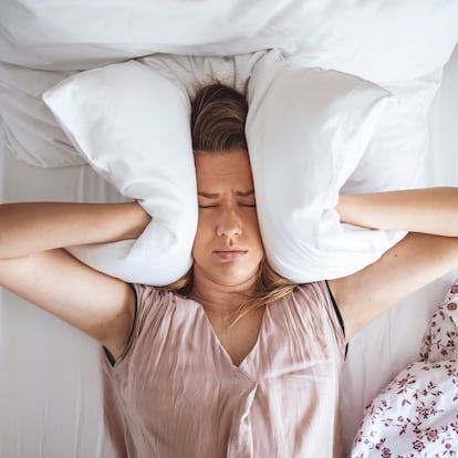 Young woman trying to sleep but disturbed by noisy neighbors and covering ears with pillows. I can's sleep with all that noise! Sleep problems. Angry woman disturbed with a noise trying to sleep