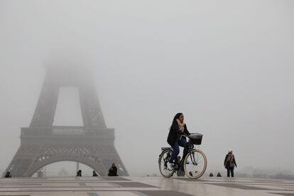 Una mujer monta una bicicleta frente a la torre Eiffel parcialmente oculta por la niebla.