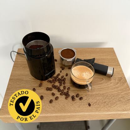 Un molinillo con gramos de café y una taza encima de una mesa.