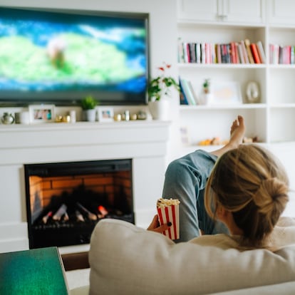 Blond woman with hair bun watching TV on a cozy sofa against fireplace indoors