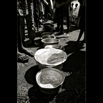 Cazuelas que se utilizan para guardar una de las dos comidas cocinadas que se ofrecen en el Centro de Tránsito. Los refugiados presentan su tarjeta de identificación y así pueden recibir una ración de gachas, frijoles y arroz. Aunque sólo se ofrecen dos comidas al día, las colas tienden a ser tranquilas y la gente espera pacientemente a obtener su plato. Otra foto durante el reparto de las gachas. Muchos niños esperan junto a los adultos, en la misma cola, para recoger sus porciones. Transportar la avena caliente en recipientes sobre la cabeza ha llevado a que algunos porteadores sufran escaldaduras y quemaduras cuando la papilla chapotea y se derrama fuera del recipiente, pero estos accidentes son relativamente escasos para la cantidad que se trae y se lleva a diario.