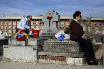 Un hombre limpia la lápida de un familiar junto a una mujer sentada en el cementerio de La Almudena de Madrid.
