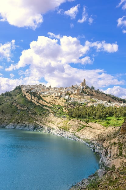 Iznájar (Córdoba). Otra fortaleza árabe dio origen y nombre a este pueblo; derivando del sustantivo ‘hisn’ (castillo) y del adjetivo ‘al-ashar’ (alegre). La población está en un lugar extraordinario: sobre una escarpada cresta de las Sierras Subbéticas, rodeada por las aguas represadas, color mar tropical, del río Genil, que forman el mayor embalse del sur de España. En la parte alta, junto a la iglesia y el castillo, se halla el Patio de las Comedias, una placita repleta de macetas azules desde donde se ve como Dios el pueblo blanco y el llamado “lago de Andalucía”. Ahí al lado, en la calle Villa, se encuentra el ascensor que usan para bajar los féretros al cementerio, porque es tan empinada la ladera que, si no, no hay manera. ¡Cosas veredes, Sancho!: un pueblo en el que los muertos, en lugar de subir, caen del cielo.