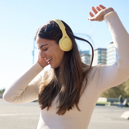 Happy young woman enjoys music with yellow wireless headphones, dancing in a sunny urban square.