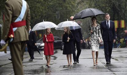 Sofía, de rojo, y Leonor, de azul marino, caminan con paraguas transparentes junto a la Reina, vestida en tonos blanco y negro.