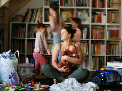 Tired, burnt-out mother with baby in her arms and out of focus children run around. Mother is sitting on floor in living room, among vacuum cleaner, laundry and children running around.