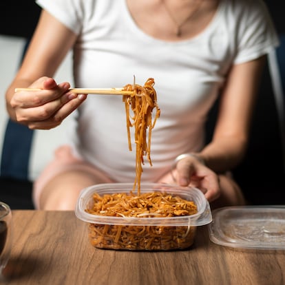 Woman having takeaway or delivery Pad Thai fried noodles with chicken meal from a plastic package with juice at home