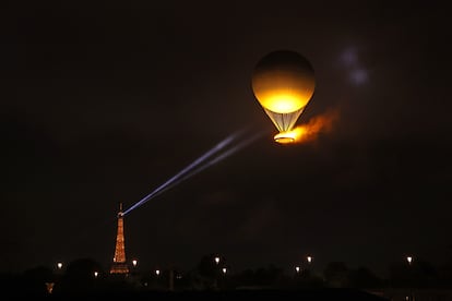 El foco de la Torre Eiffel ilumina el globo aerostático con el pebetero olímpico por el cielo de París.