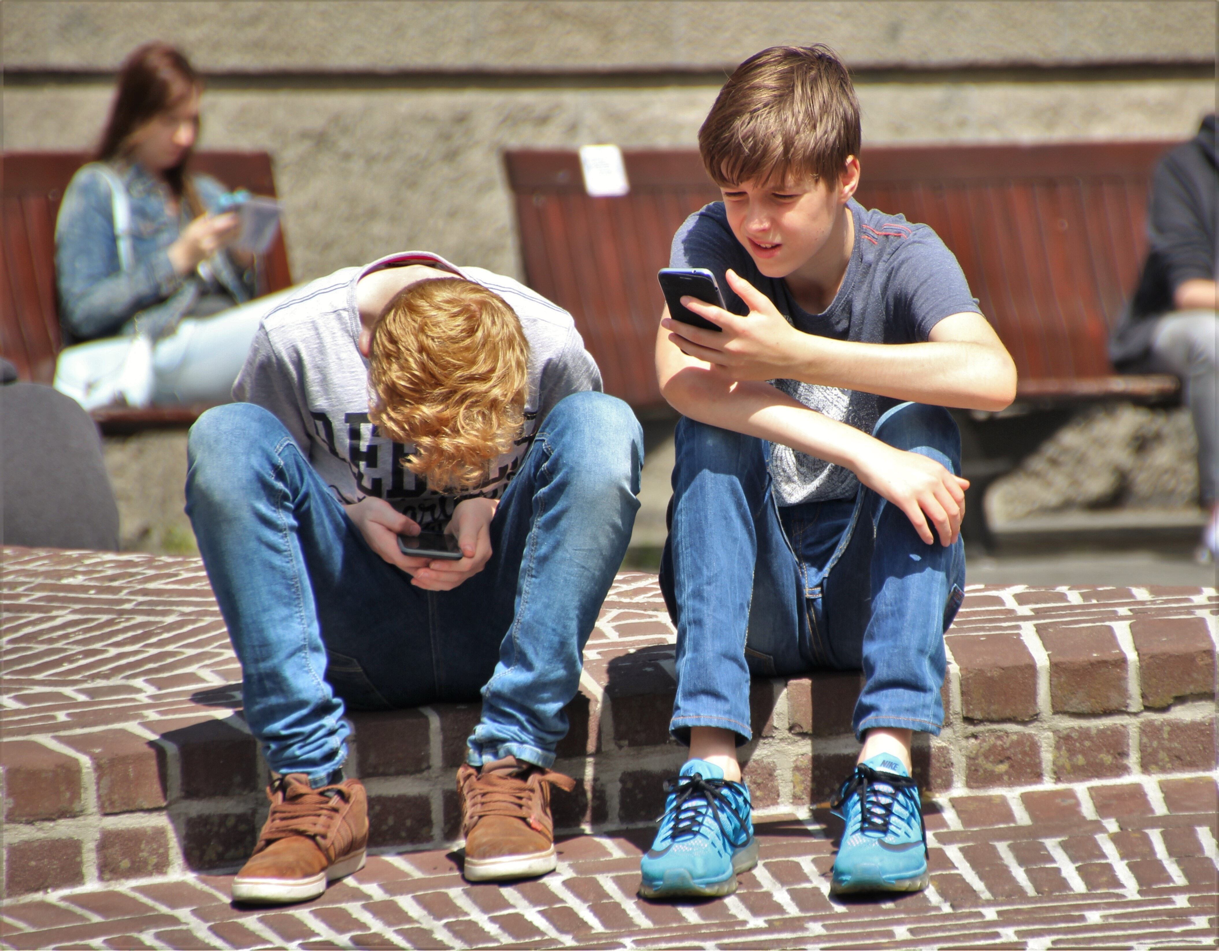 Un par de adolescentes miran el móvil, sentados en un bordillo.