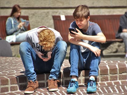 Un par de adolescentes miran el móvil, sentados en un bordillo.