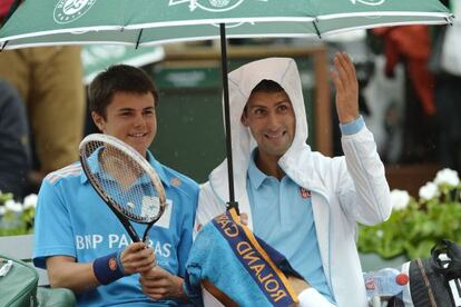 Novak Djokovic bromea con un recogepelotas al que guarece de la lluvia.