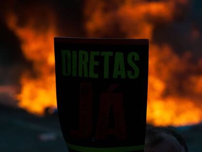 Manifestante com um cartaz em defesa de eleições antecipadas diretas, em Brasília. Ao fundo, barricada com fogo na Esplanada dos Ministérios.