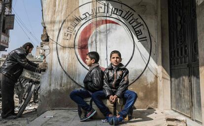 Niños sirios se sientan en una maleta frente a un mural de la Luna Roja Siria en Duma (Siria).