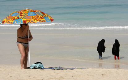 Una turista coloca una sombrilla mientras dos mujeres se mojan los pies en una playa de Dub&aacute;i.