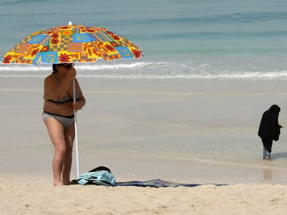 Una turista coloca una sombrilla mientras dos mujeres se mojan los pies en una playa de Dub&aacute;i.