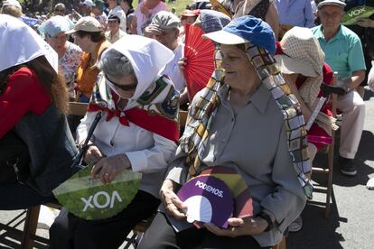 Dos Mujeres con abanicos de Vox y Podemos asisten a la misa en la pradera.