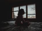 Little girl sits on a bed in a dark bedroom with blue sky and trees can be seen behind her in the window.