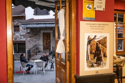 Dos vecinos de Somiedo toman una cerveza en una terraza del pueblo. A la derecha, cartel que anuncia una exposición sobre osos.
