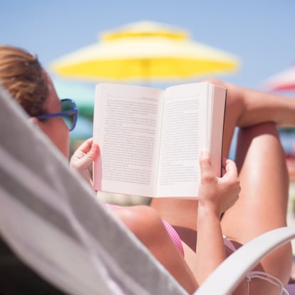 A person relaxes on a beach chair, engrossed in a book while basking in sunlight. Vibrant umbrellas provide shade against the clear blue sky, creating a serene atmosphere.