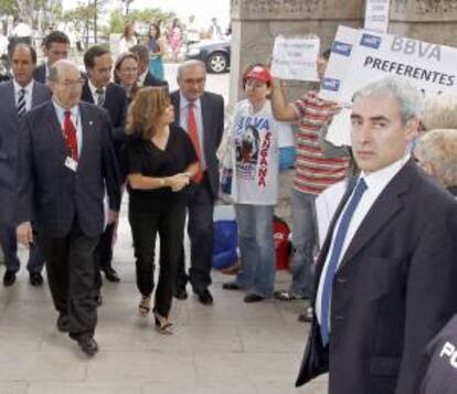 La vicepresidente del Gobierno, Soraya Sáenz de Santamaría (2i), observa la cartelería que portan afectados por las participaciones preferentes, a su llegada al palacio de La Magdalena, para intervenir en el seminario: Leyes sabias y justas. Una apuesta por la calidad de las leyes" en la Universidad Internacional Menéndez Pelayo.