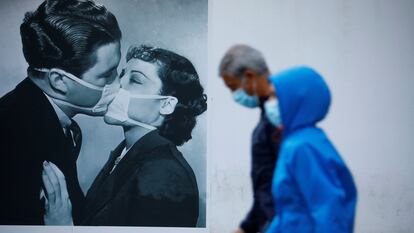 Unas personas protegidas con mascarilla pasan junto a una foto en la pared de un beso con mascarillas en Córdoba.