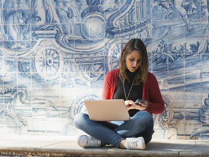 Una mujer trabaja con su móvil y portátil en una calle de Lisboa, con los tradicionales azulejos a su espalda.