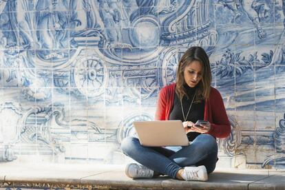 Una mujer trabaja con su móvil y portátil en una calle de Lisboa, con los tradicionales azulejos a su espalda.