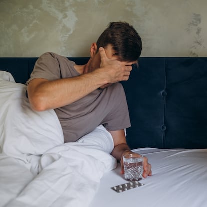 man in bed holds his hand on his head with a headache and drinks pills