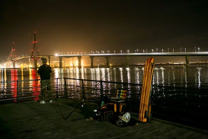 Un pescador nocturno observa el nuevo puente, al fondo, ya iluminado.