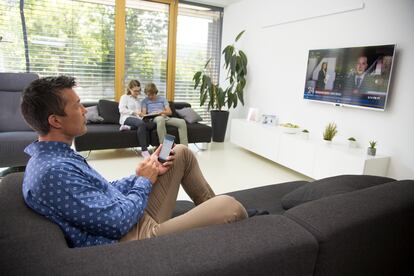 Un hombre ve la televisión mientras usa su teléfono móvil y sus hijos la tableta.