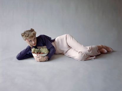 Model, lying on her side, holding her head in one hand, with other arm around bowl of grapes and cherries, and wearing a short blue-purple wool jacket and pale pink one-piece pajamas by Martini, with Revlon's Orchids To You nail enamel and lipstick. (Photo by?Richard Rutledge/Cond Nast via Getty Images)