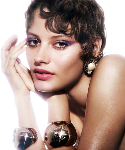 Para rasgar la mirada se dibuja una forma triangular siguiendo la línea baja de las pestañas, con el morado de la paleta Baroque Les 4 Ombres Boutons. En los labios, Rouge Allure Velvet (tono Élégante). Pendientes con perlas y cadenas y brazaletes metálicos. Todo de CHANEL.