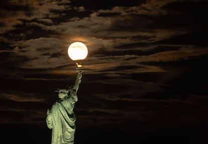 La superluna del Esturión aparece entre nubes detrás de la Estatua de la Libertad en la ciudad de Nueva York, vista desde Jersey, Nueva Jersey.