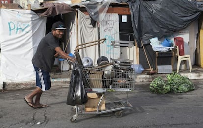 Homem empurra carrinho de supermercado repleto de objetos de metal. Atrás, barracos improvisados onde dormem os sem-teto que vivem na zona.