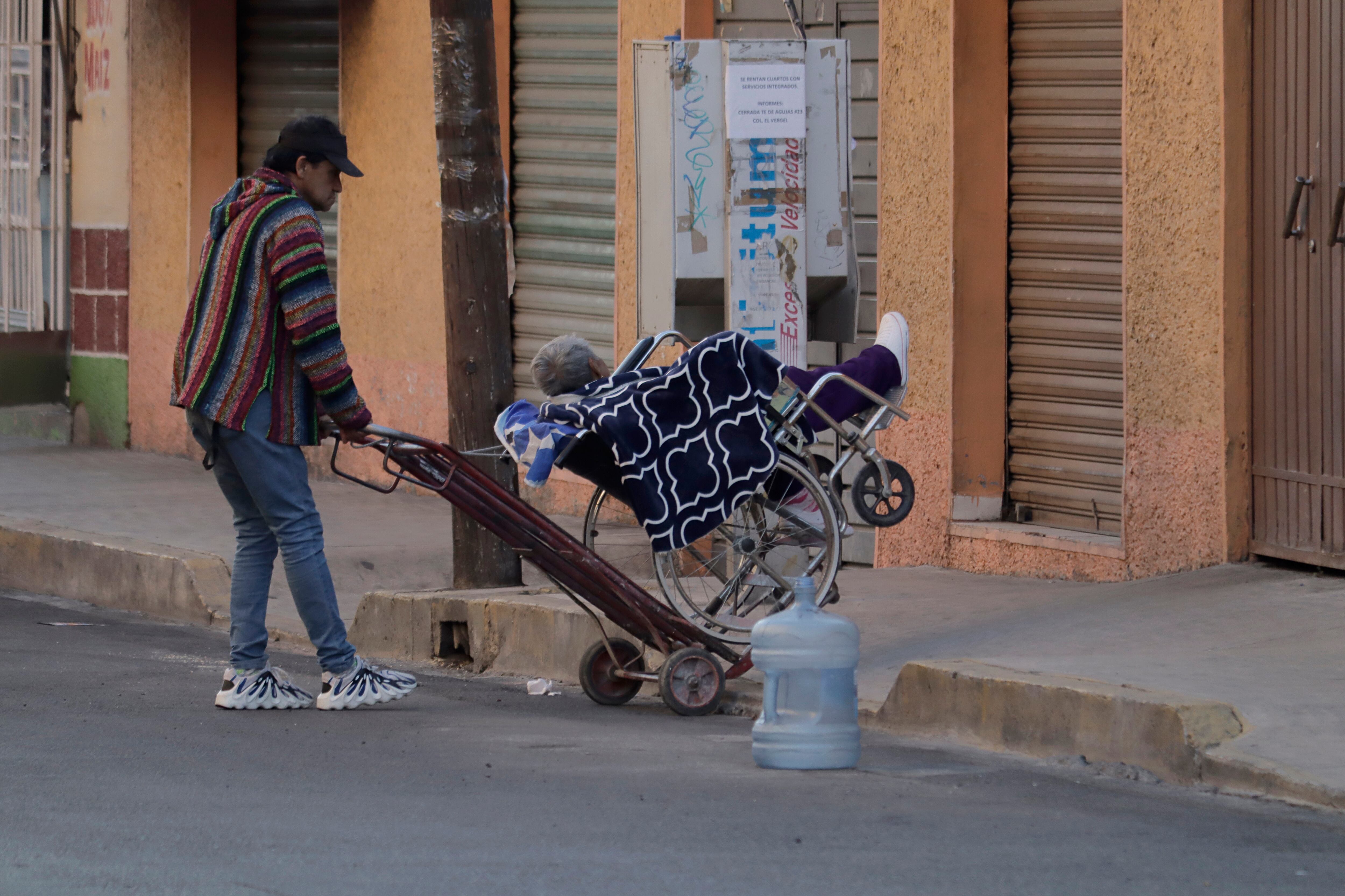 Un hombre ayuda a una adulta mayor en silla de ruedas a subir a una acera utilizando una carretilla, en Ciudad de México.