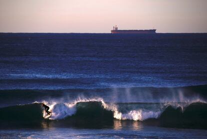 Un barco que espera una carga de carbón se puede ver detrás de un surfista montando una ola en Merewether Beach, en Newcastle, ubicado al norte de Sidney (Australia).
