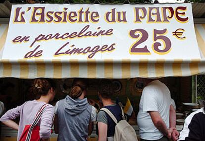 Varios turistas curiosean un puesto en Lourdes que anuncia la venta del plato del Papa en porcelana de Limoges.