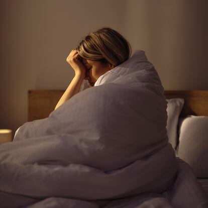 Young woman holding her head in pain while being wrapped in a duvet on a bed in bedroom. Photographed in medium format.