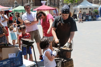 La forjadora artística Inma López enseña a Carlota, de tres años, a utilizar el yunque y el martillo en la feria. Juntas, golpean una flor de hierro.