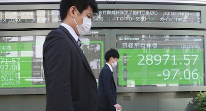 Dos personas caminan enfrente de un panel de un bróker japonés.