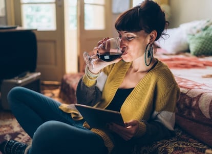 Una chica bebe una copa de vino mientras usa la tableta. GETTY