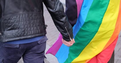 Casal de mãos dadas diante da bandeira multicolorida que identifica a causa LGBTI.